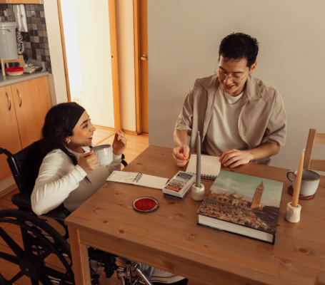 Una persona en silla de ruedas y una en silla sin ruedas sentadas en el comedor de su casa